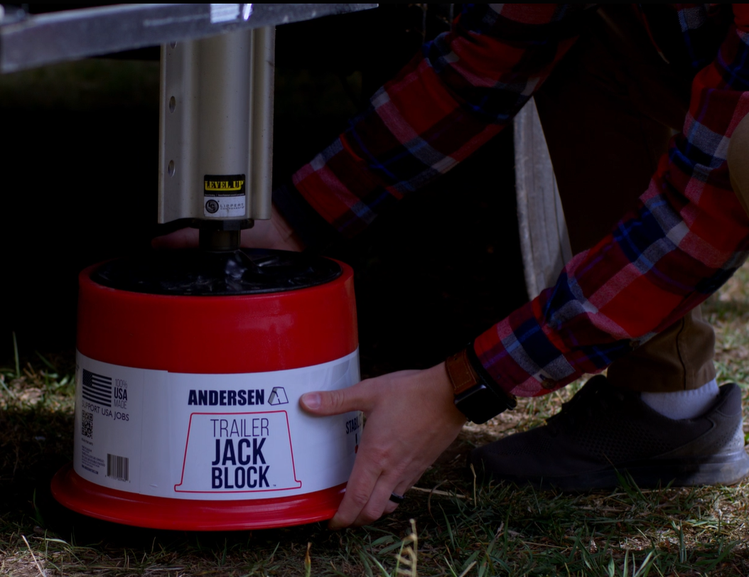 A man placing a red and white Andersen Trailer Jack Block under a fifth-wheel trailer stabilizer. The video highlights the product’s 90% stability increase, magnetic leveling system, and lifetime warranty.